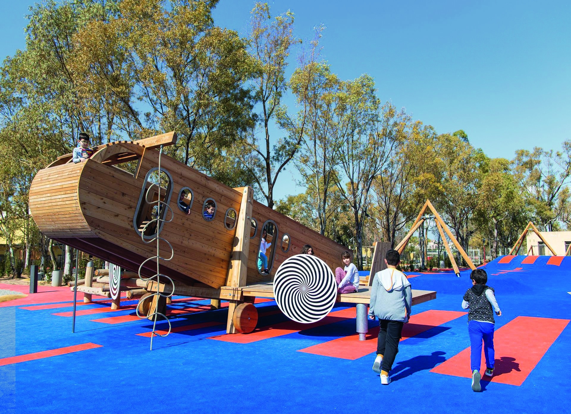 Mehrere Kinder spielen im Freien auf einem großen Holzflugzeug auf einem blau-orangenen Boden.