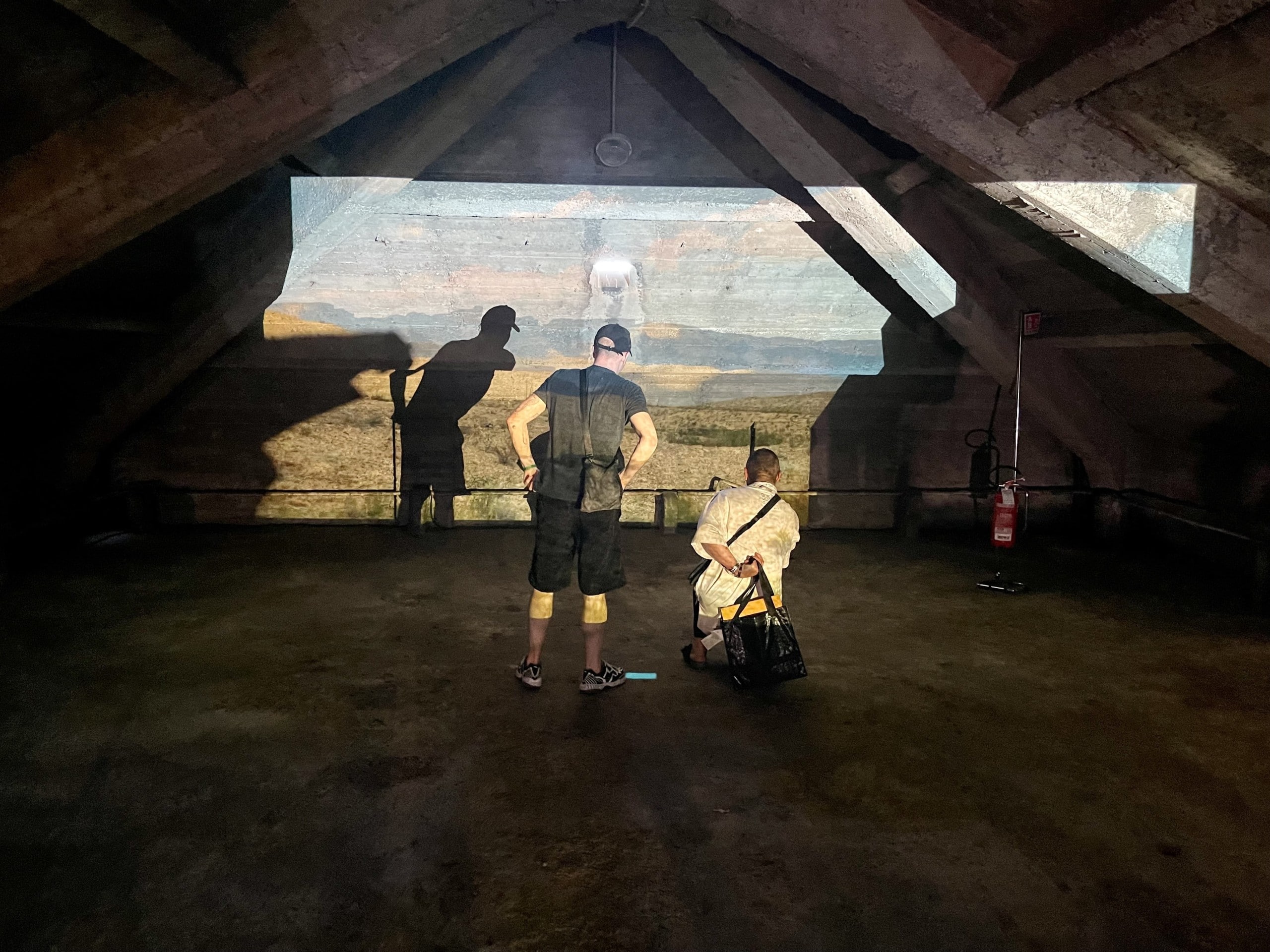 Blick auf eine Projektion in einem alten Dachraum, vor der eine Person steht und eine Person kniet. Alcova 2022, Foto: Baumeister/Fabian Peters