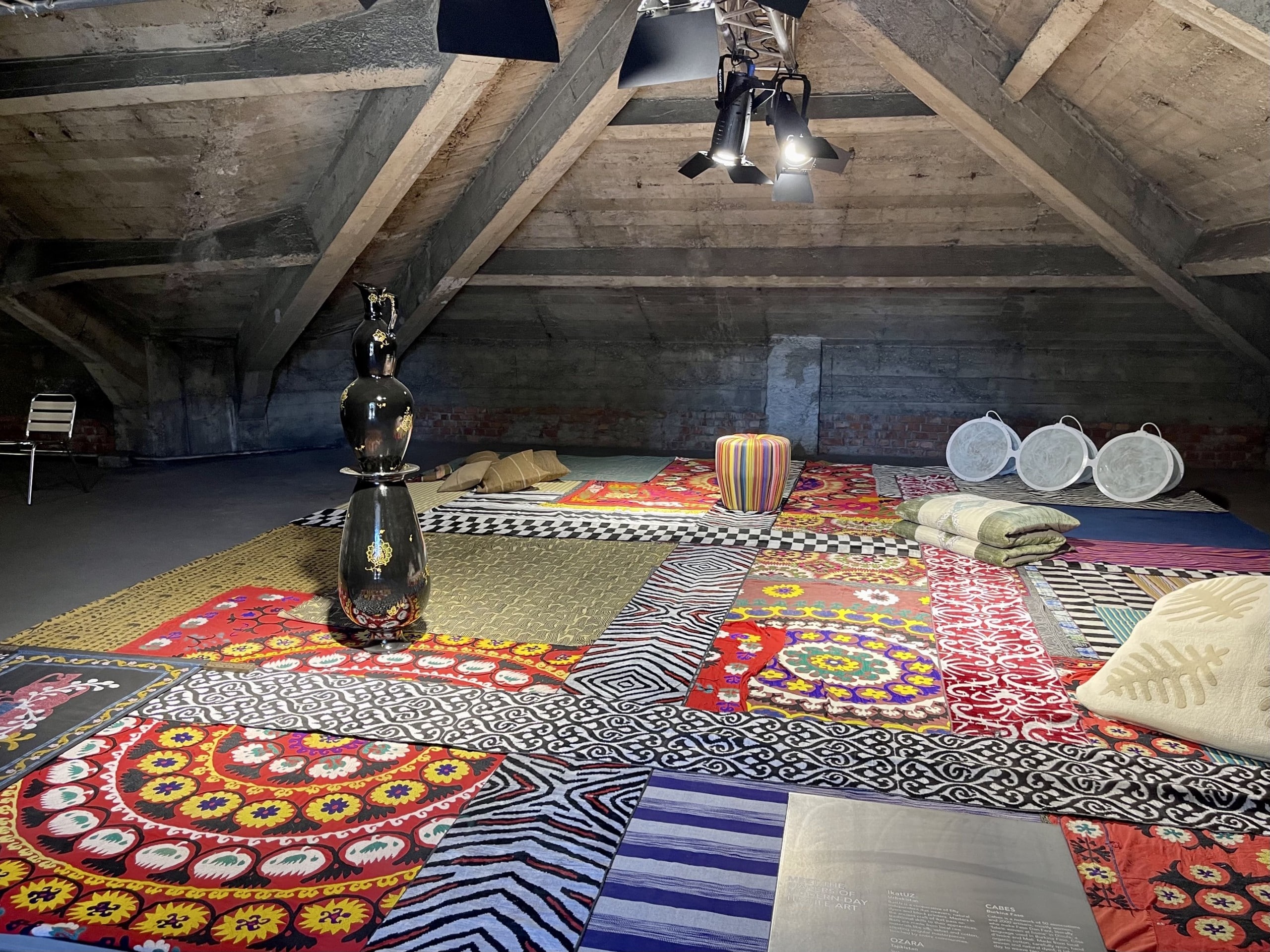 Blick auf mehrere Bunte Teppiche auf dem Boden eines Dachbodens, auf dem ein großes schwarzes Keramikgefäß steht. Alcova 2022, Foto: Baumeister/Fabian Peters