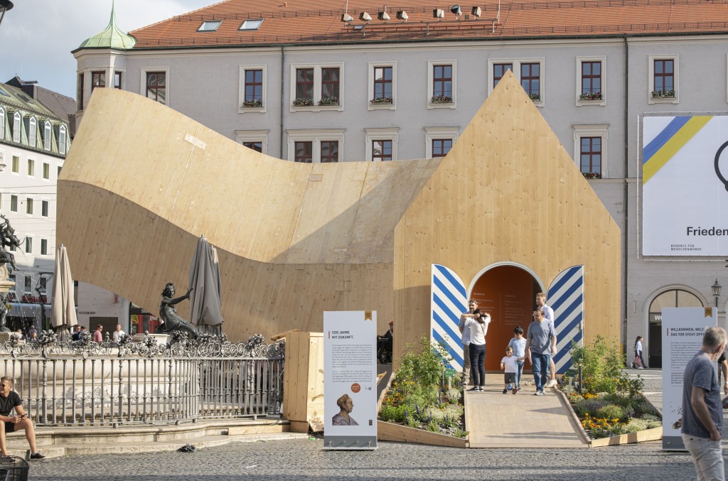Blick auf einen länglichen, sich gekurvt schlängelnden Holzpavillon vor einem Gebäude in Augsburg anlässlich des Fests „500 Jahre Fuggerei“. MVRDV, NEXT500 Pavillon, Augsburg, Foto: © Saskia Wehler