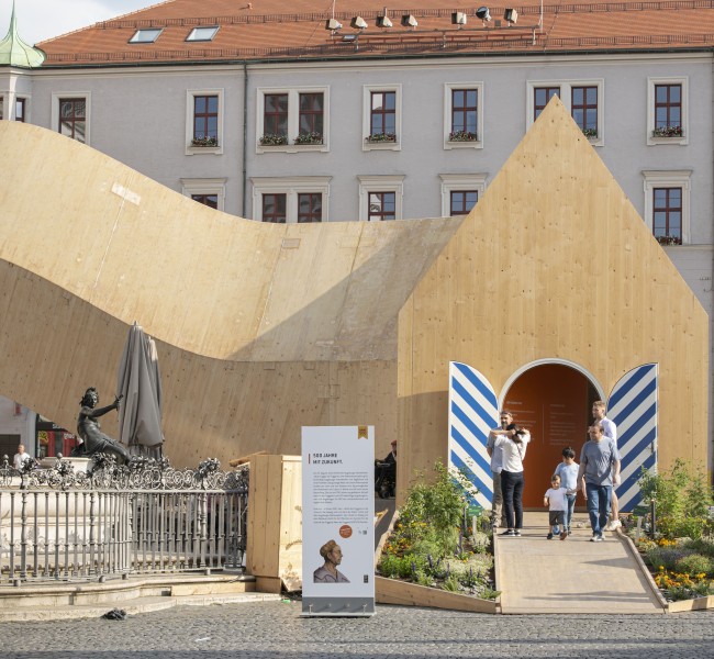 Blick auf einen länglichen, sich gekurvt schlängelnden Holzpavillon vor einem Gebäude in Augsburg anlässlich des Fests „500 Jahre Fuggerei“. MVRDV, NEXT500 Pavillon, Augsburg, Foto: © Saskia Wehler