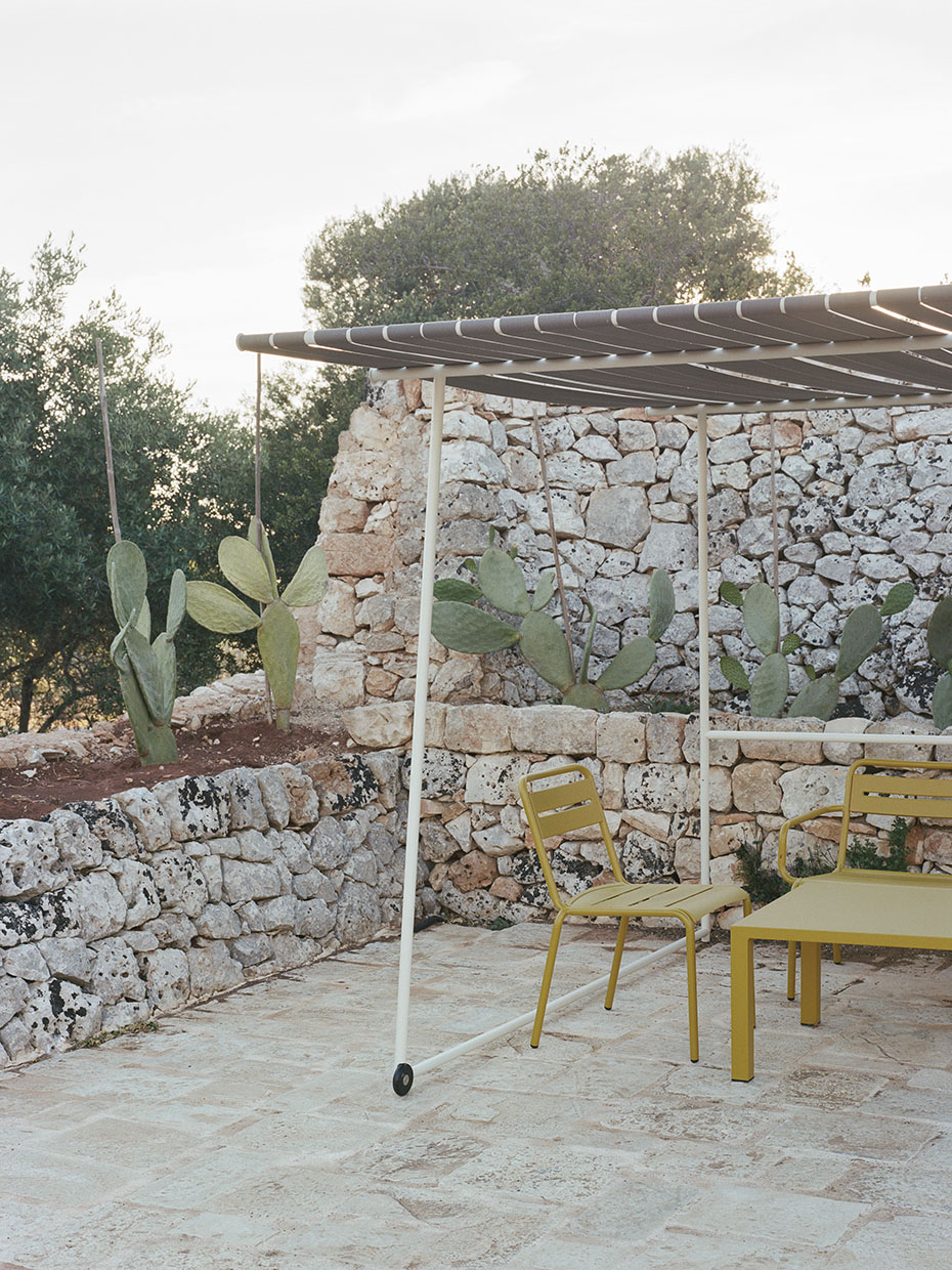 Blick auf eine steinerne Mauer, davor ein Gestell mit Sonnenschutz, darunter ein gelber Tisch mit Stühlen, daneben Kaktuspflanzen. Valari, Masseria Belvedere, Carovigno, Foto: ©Lorenzo Zandri