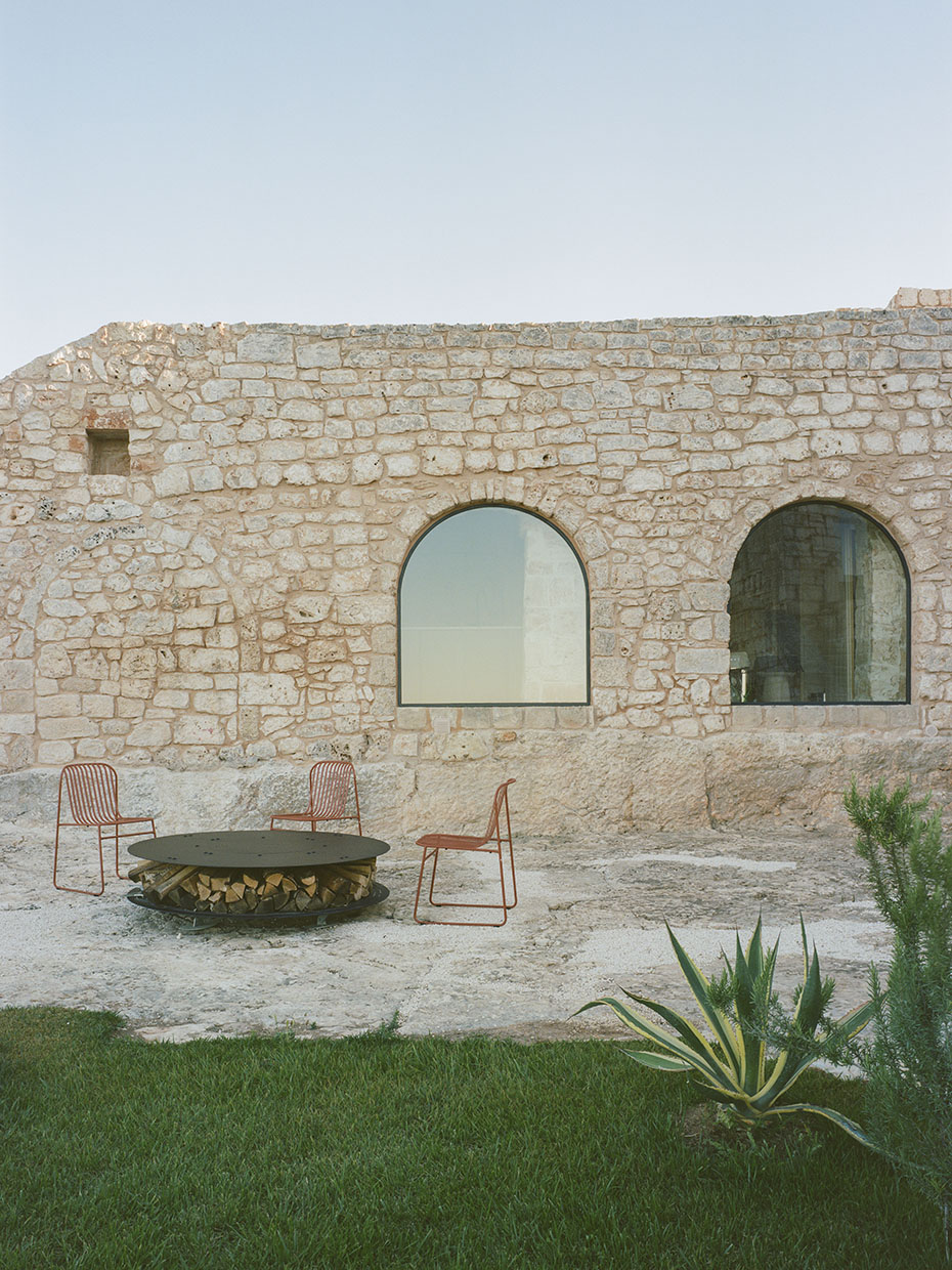 Blick auf die Außenwand eines Gebäudes aus hellgelbem Stein, davor eine Feuerschale und drei rote Stühle. Valari, Masseria Belvedere, Carovigno, Foto: ©Lorenzo Zandri