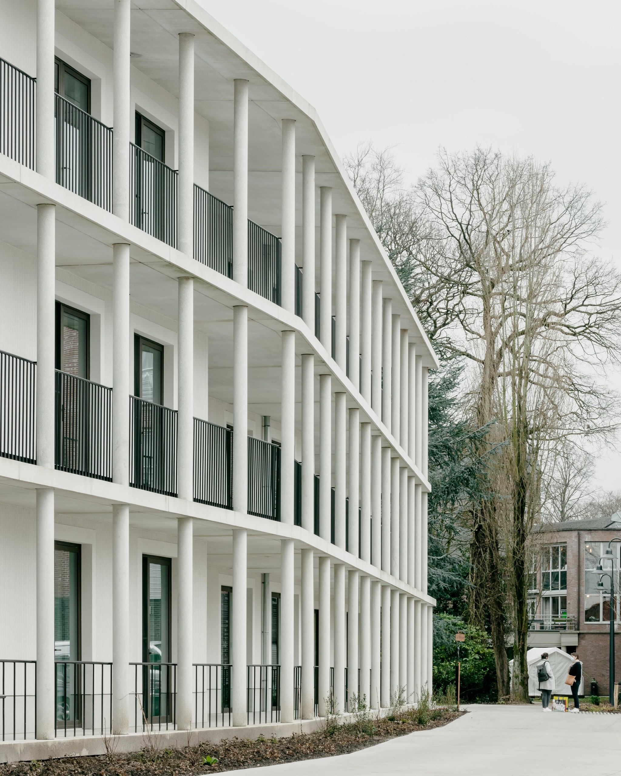 Schräger Blick auf die Fassade eines dreistöckigen, weißen Gebäudes mit vorgelagerten Balkonen auf der gesamten Breite des Gebäudes, dem Neubau, der an die Villa Landhuis in Kortrijk angeschlossen ist. Stijn Bollaert