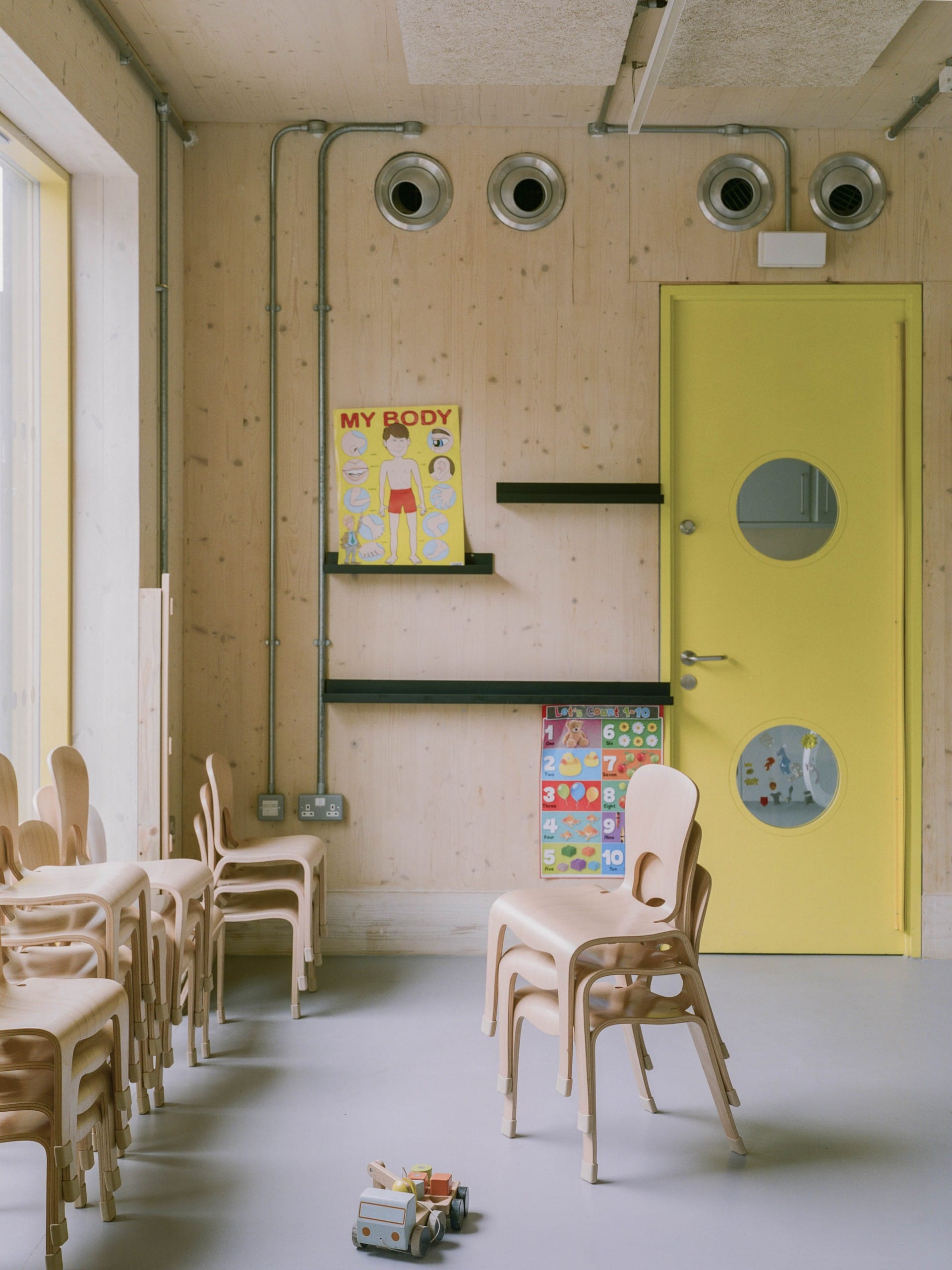 Ausschnitt eines Innenraums, Wand aus hellem Holz, angeschnitten eine gelbe Türe, im Vordergrund mehrere gestapelte Holzstühle. Foto: Lorenzo Zandri