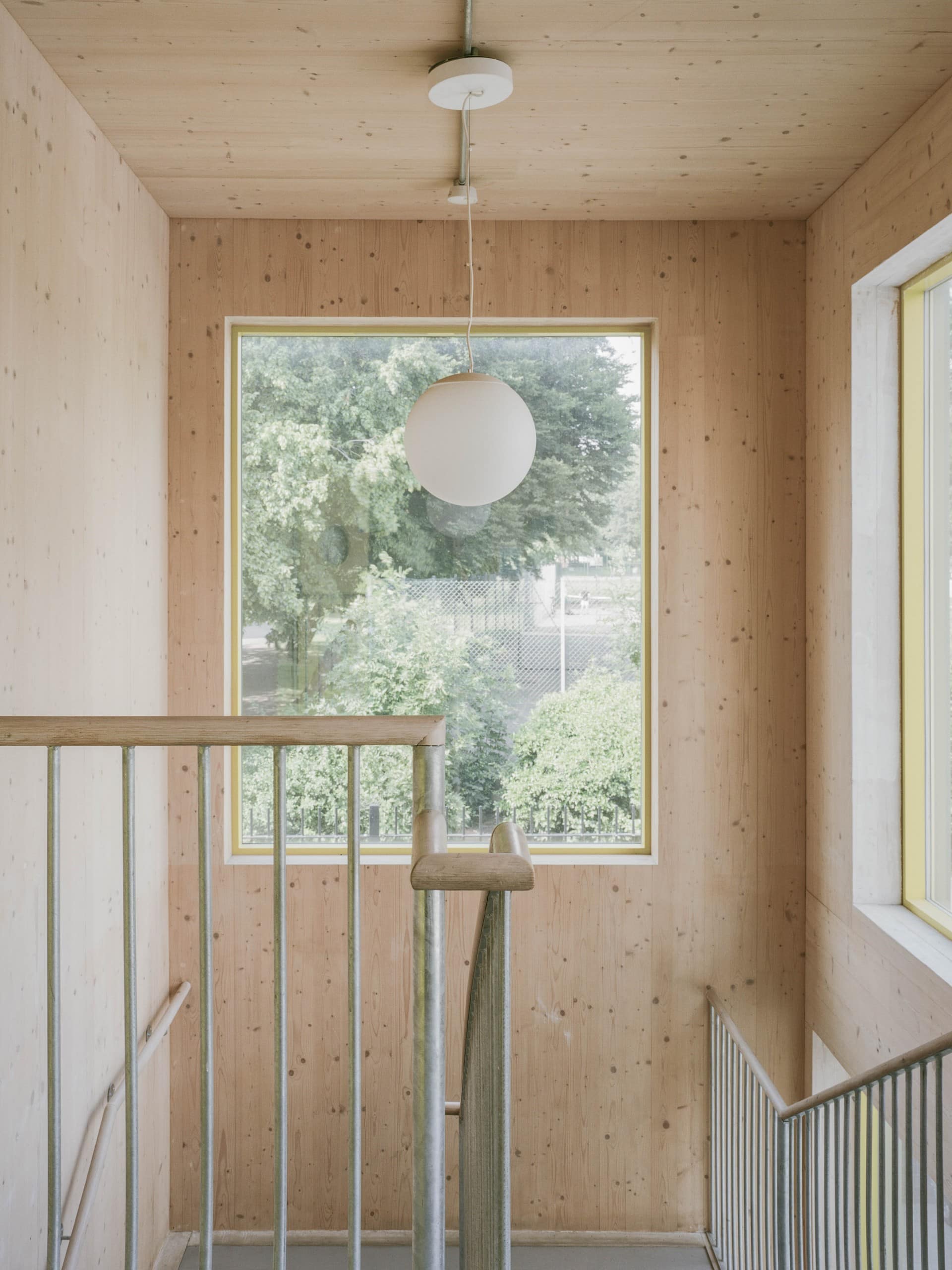 Blick in ein Treppenhaus, Wände und Decke aus hellem Holz, metallene Treppengeländer mit senkrechten, dünnen Stäben, große Fenster in den Wänden, von der Decke hängt eine kugelförmige, weiße Lampe herunter. Foto: Lorenzo Zandri