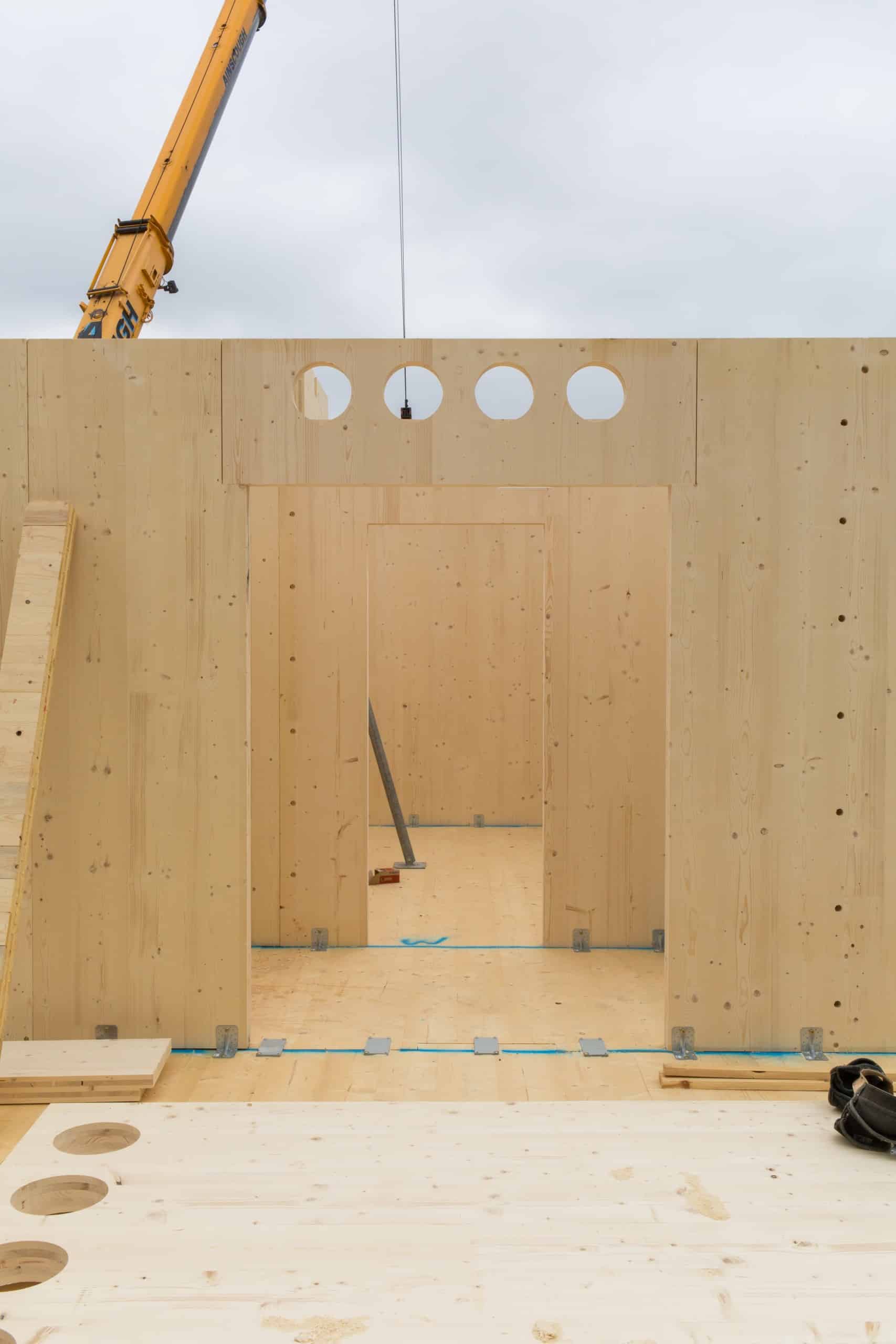 Errichtung eines Gebäudes in Holzbauweise, Blick auf Wand, die gerade aufgestellt wird, im Hintergrund ist ein Kran angeschnitten. Dyvik Kahlen Architects, Kindergarten und Gemeindezentrum, London, Foto: Antoine Espinasseau