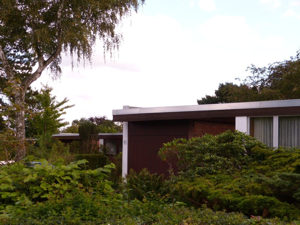 Das Bild zeigt einen eingeschossigen Bungalow mit Glasfassade. Er steht hinter Hecken und von Bäumen umgeben.