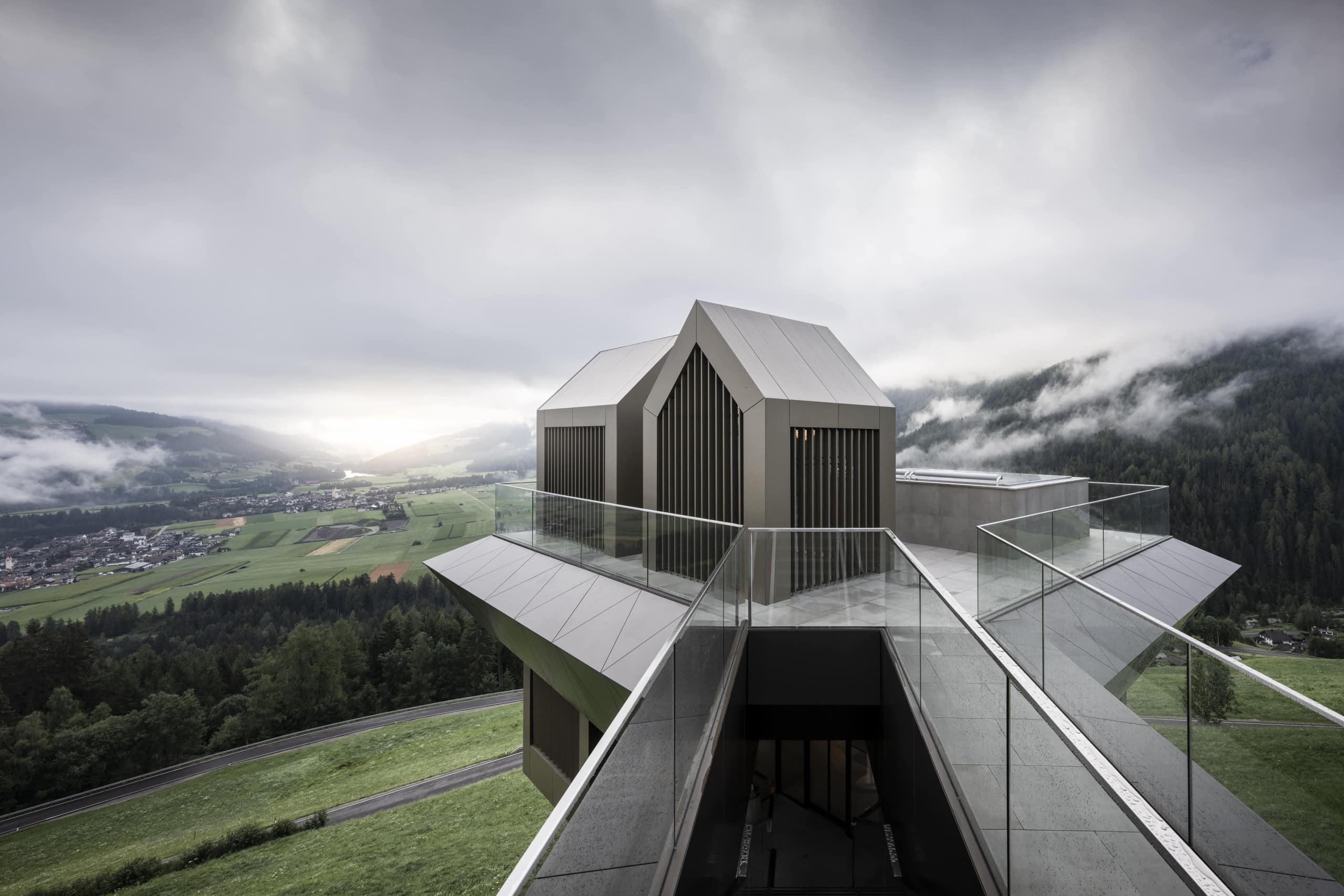 Blick auf eine Plattform mit mehreren kleinen Hütten mit Aluminiumfassade, im Hintergrund Berge, Hügel und graue Wolken. Noa* Network of Architecture, Hub of Huts, Hotel Hubertus Olang, Foto: ©AlexFilz