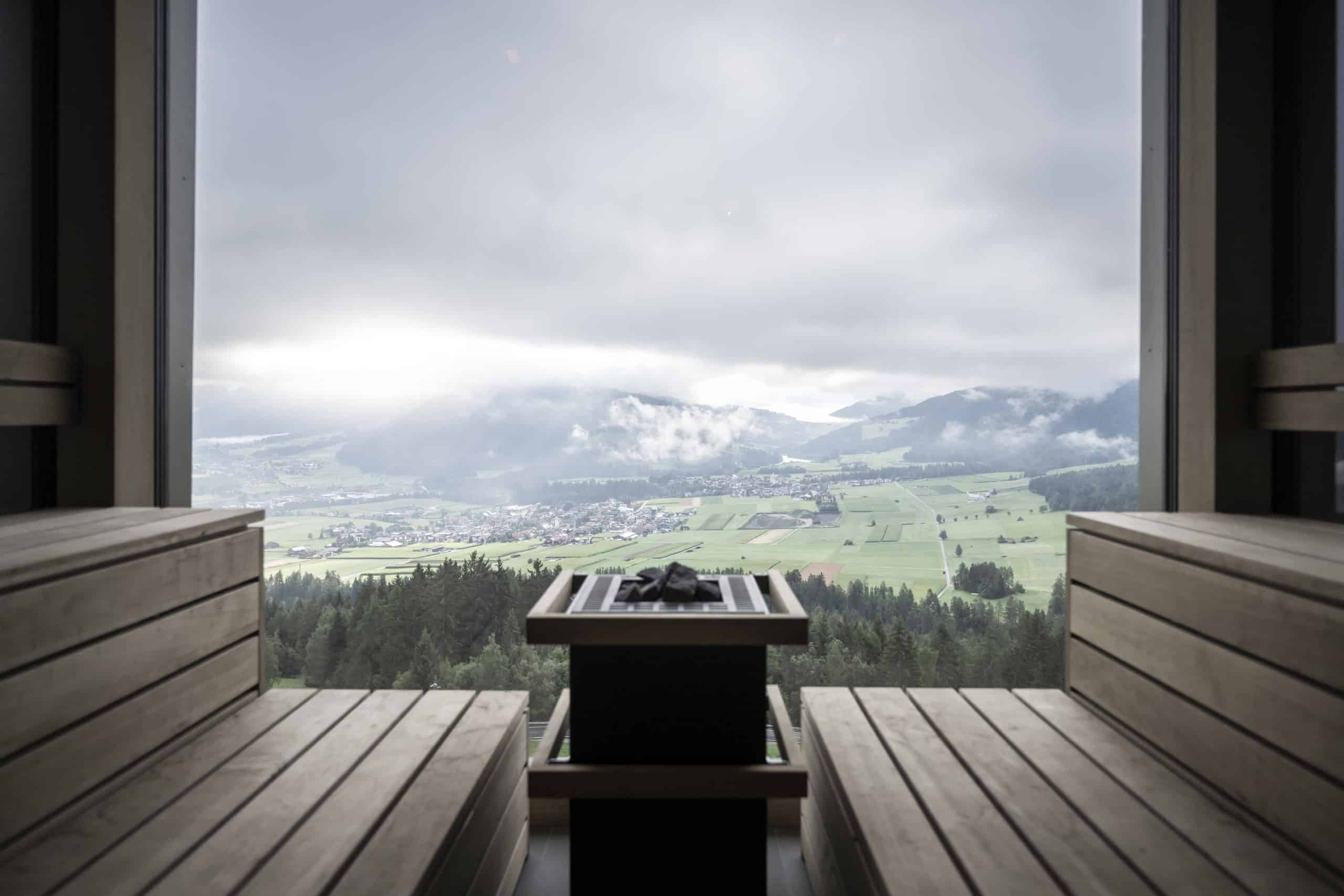 Im Vordergrund zwei hölzerne Sitzbänke einer Sauna, dazwischen ein Saunaofen, dahinter ein bodentiefes Fenster mit Ausblick auf eine weite Landschaft mit Bergen. Noa* Network of Architecture, Hub of Huts, Hotel Hubertus Olang, Foto: ©AlexFilz