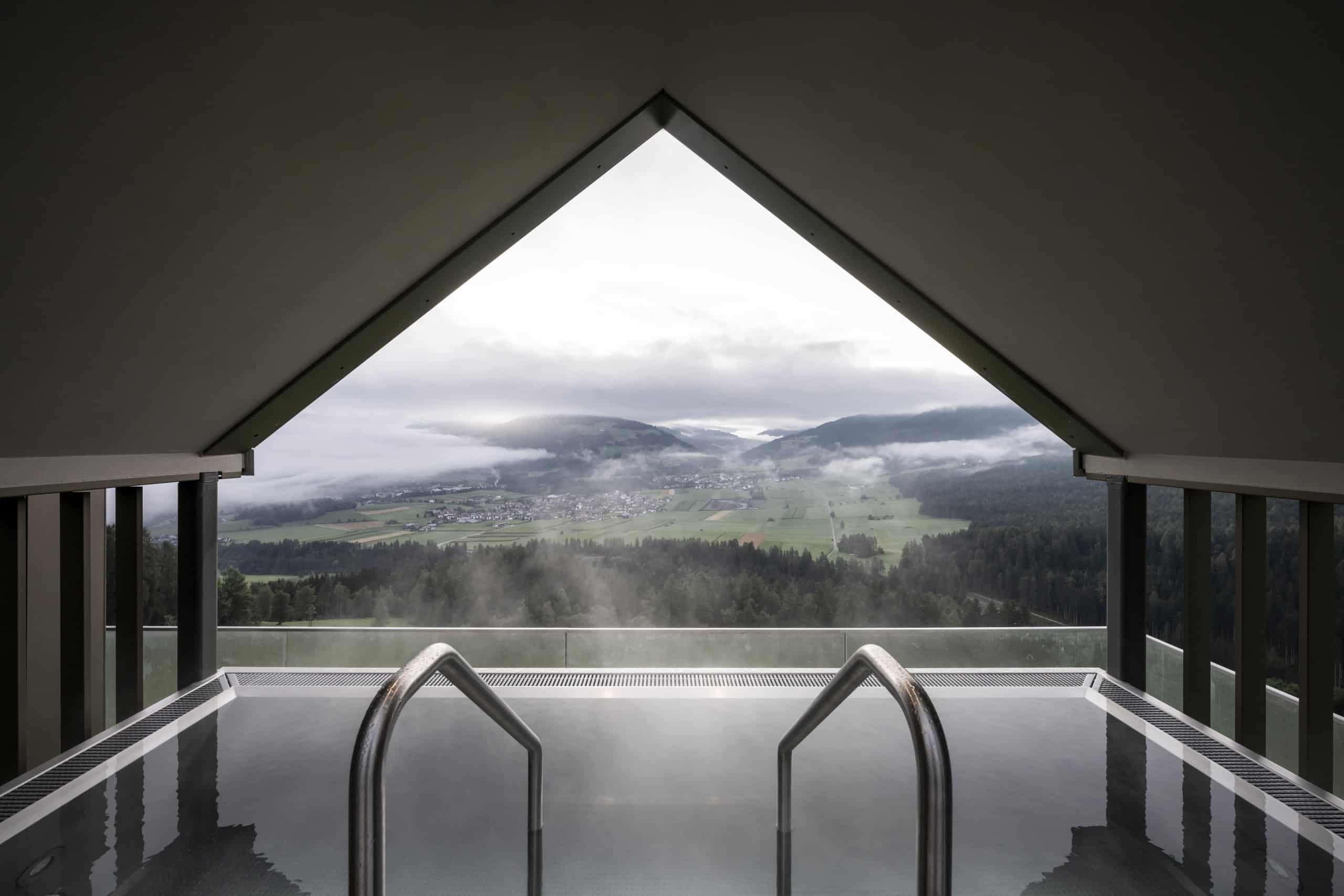 Ein Pool in einem Raum mit Satteldach, an der Stirnseite ein wandgroßes Fenster mit Ausblick auf eine Berglandschaft. Noa* Network of Architecture, Hub of Huts, Hotel Hubertus Olang, Foto: ©AlexFilz