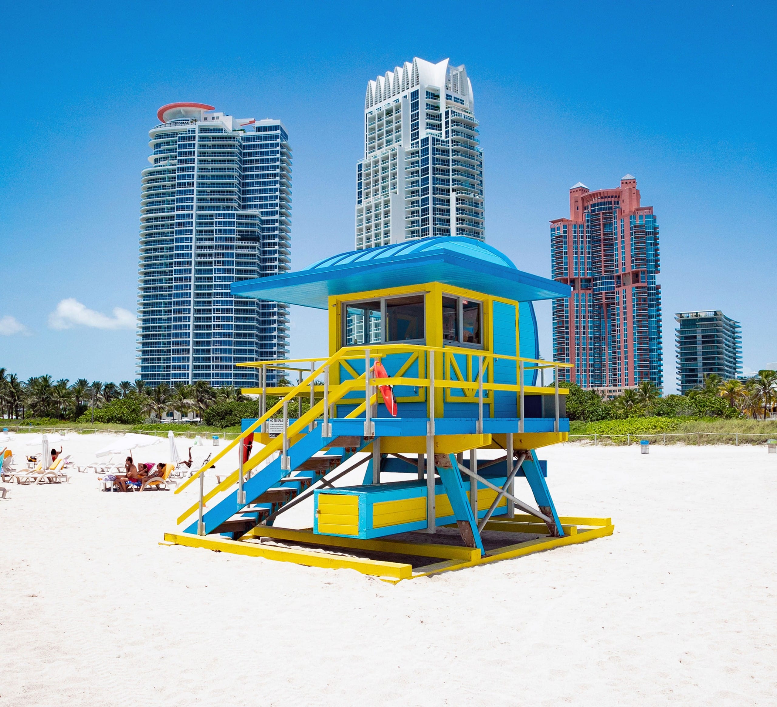 Ein hölzernes Strandwächterhäuschen in Blautönen und Gelb vor einer Skyline mit drei Hochhäusern. Miami Beach, Lifeguard Tower, William Lane, Foto: Archiv Architekten